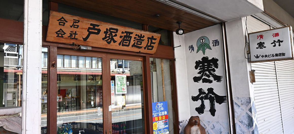 Close-up of the brewery's storefront entrance with the Totsuka Shuzoten wooden sign and the Kanchiku brand name painted on the wall.