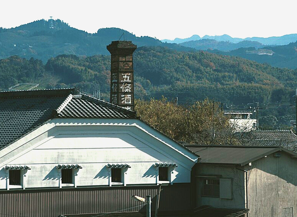 Brewery roofline and chimney marked "Goshin Shuzo" (五條酒), with surrounding hills.