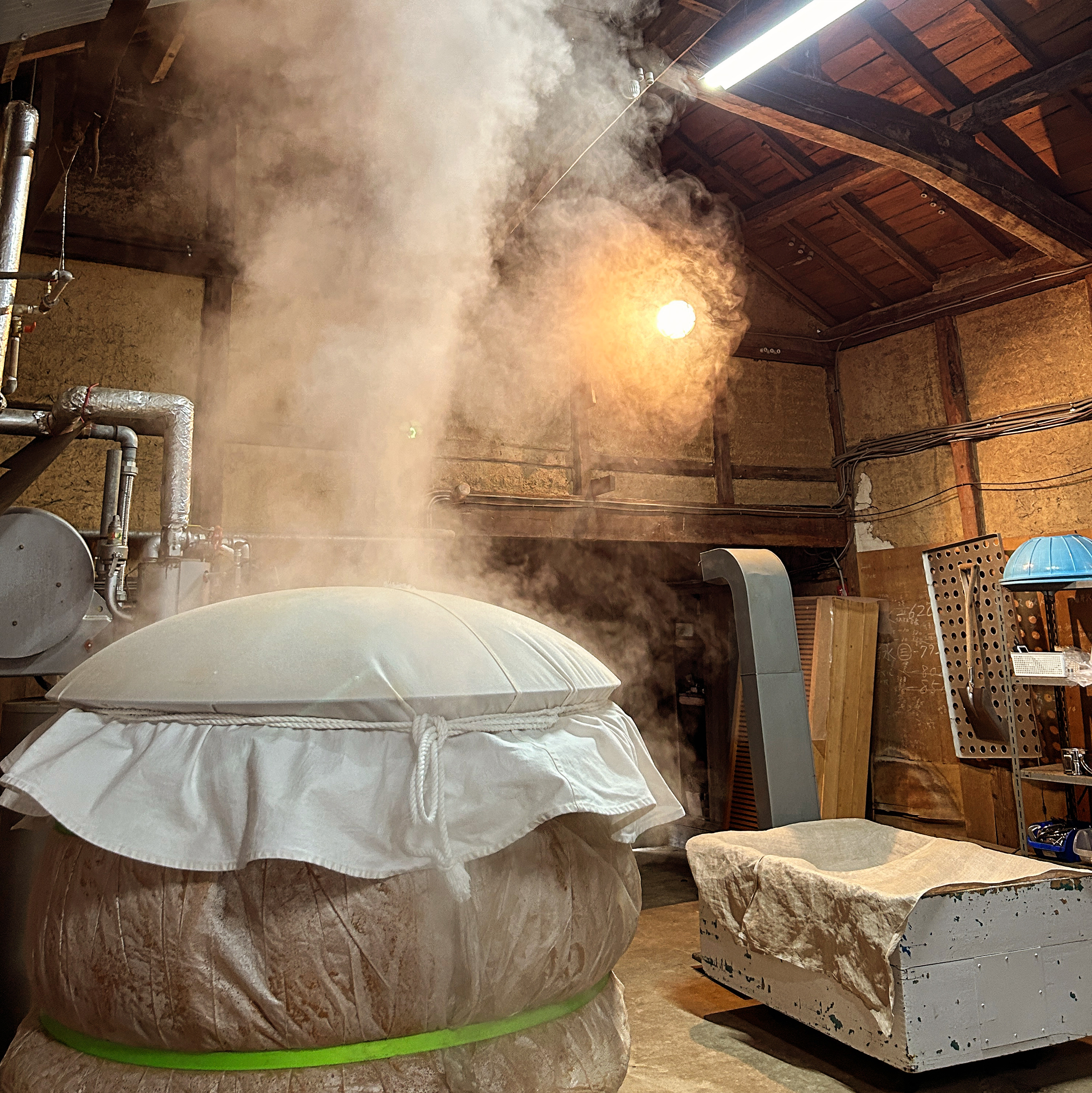 Steam rising from a fukuro-shibori pressing bag inside the brewery's wooden-beamed kura.