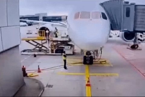 A commercial airplane parked at an airport gate with a jet bridge attached, ground crew present, and a cargo loader at the rear.