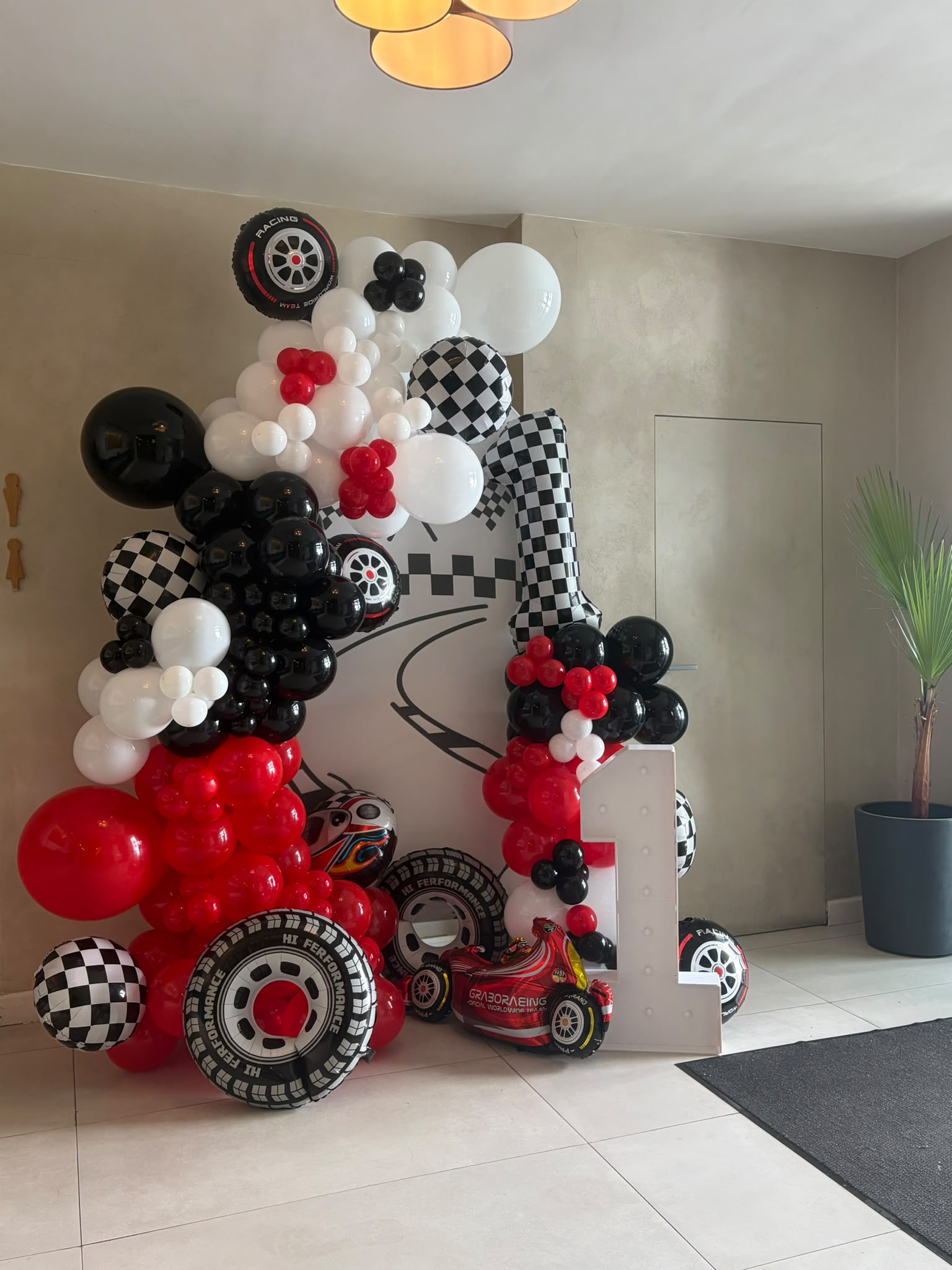 Decor de aniversare cu baloane roșii, negre și albe, panou cu pistă de curse și cifra 1