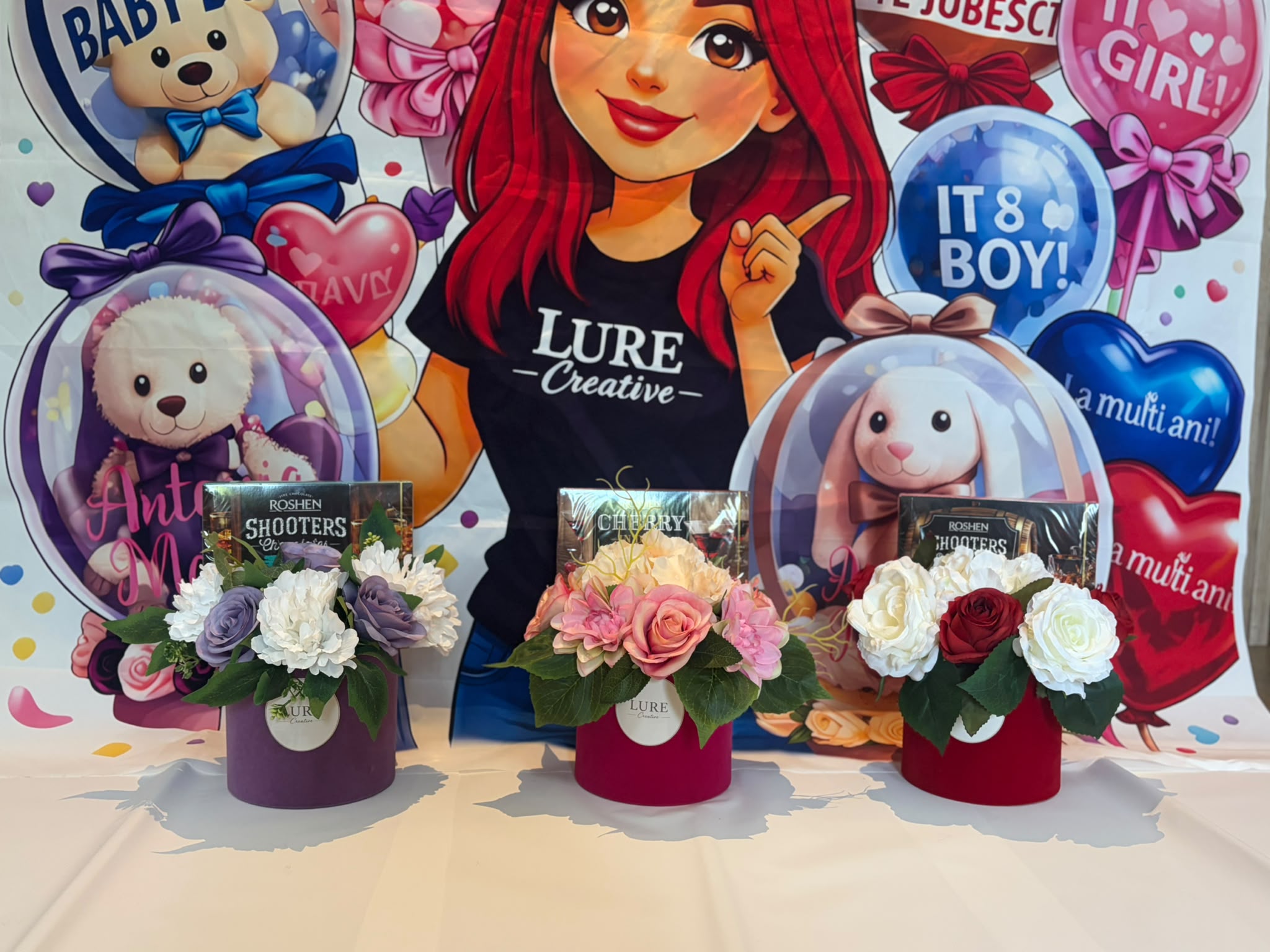 Trei aranjamente florale in cutii mov, roz si rosu alaturi de cutii cu praline de ciocolata