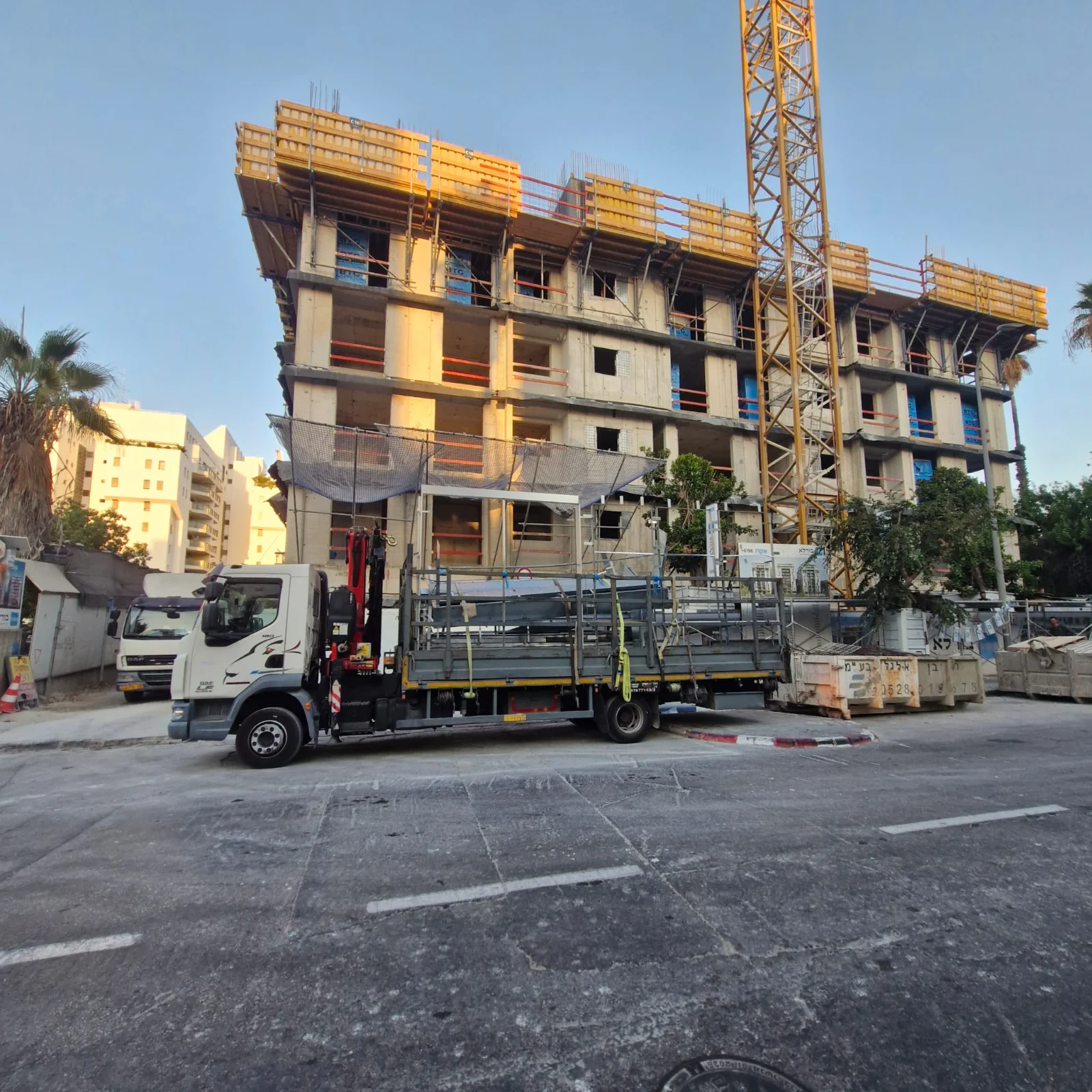 משאית מנוף טעונה חומרי בנייה מול בניין רב קומות בשלבי בנייה עם מנוף צריח