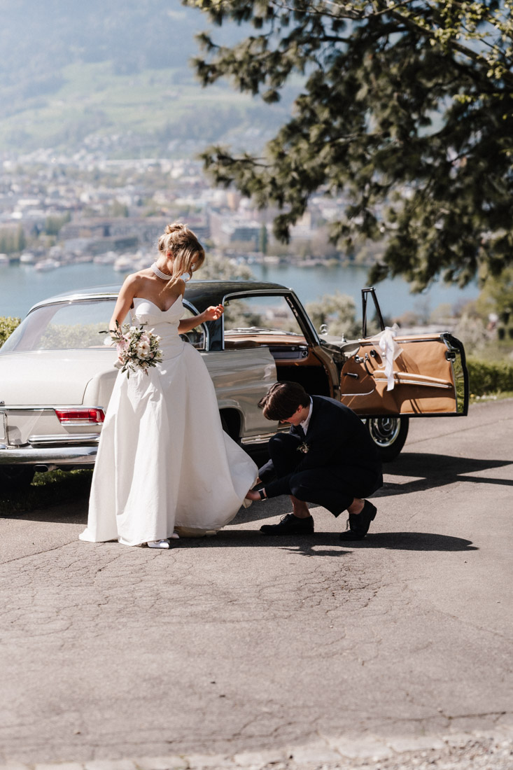 Braut im Kleid mit Blumenstrauss bei Oldtimer und Mann hilft ihr