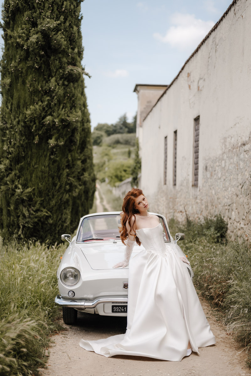 Elegante Braut steht neben klassischem Vintage-Auto auf malerischer Straße in der Toskana