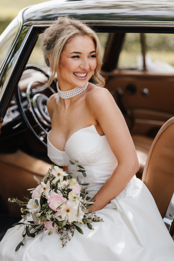 Laechelnde Frau im weissen Kleid mit Blumenstrauss und Perlenkette im Auto