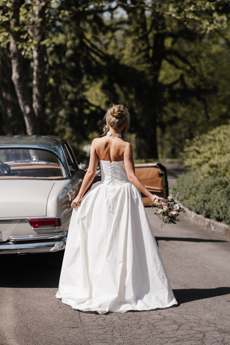 Frau im weissen Kleid mit Blumenstrauss neben Oldtimer und Bäumen