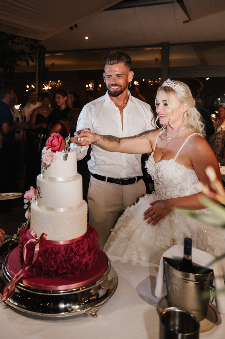 Paar schneidet Hochzeitstorte mit rosa Rosen und rotem Dekor an