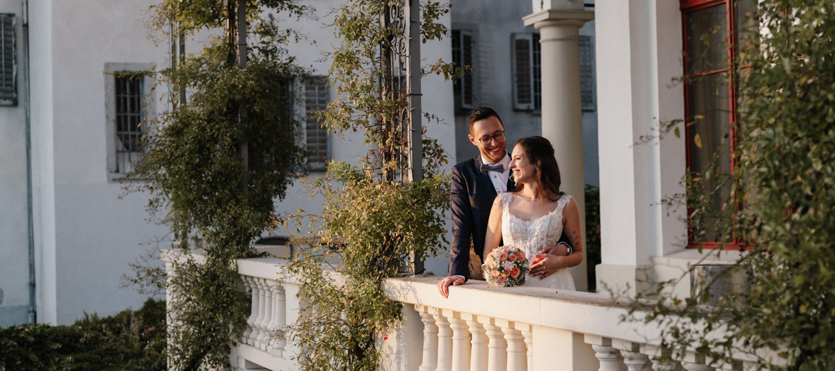 Paar lächelt auf Balkon mit Blumenstrauss und Blick zum Gebäude