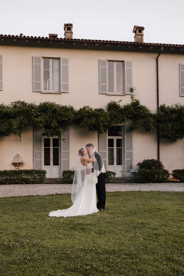 Paar küsst sich mit Schleier vor Haus mit Fensterläden und viel Grün