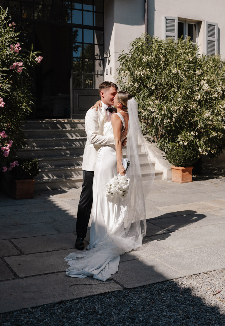 Paar umarmt sich mit Brautkleid Schleier und Blumenstrauß vor Haus