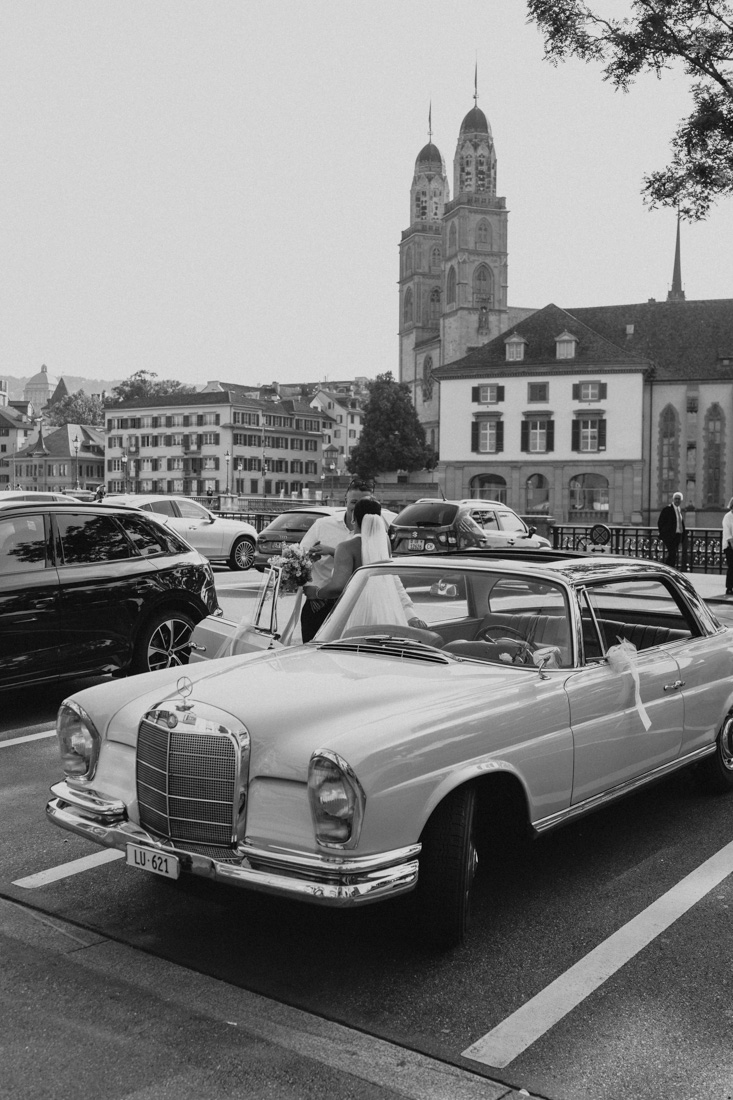 Braut mit Schleier und Bräutigam stehen vor einem Oldtimer in Zürich