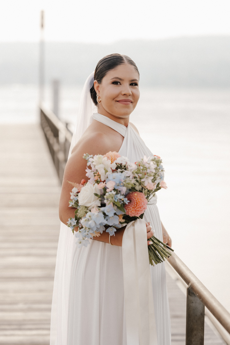 Frau im weissen Kleid mit Schleier und Blumenstrauss auf Steg am Wasser