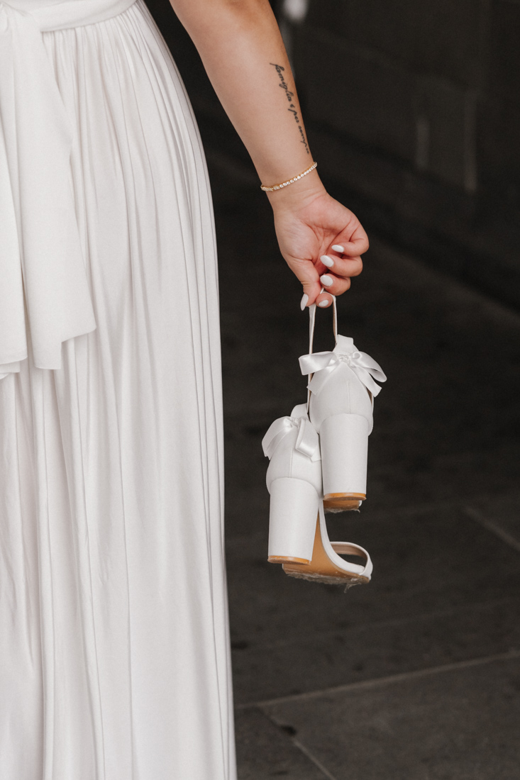 Hand hält weisse Schuhe mit Schleifen bei weissem Kleid und Tattoo