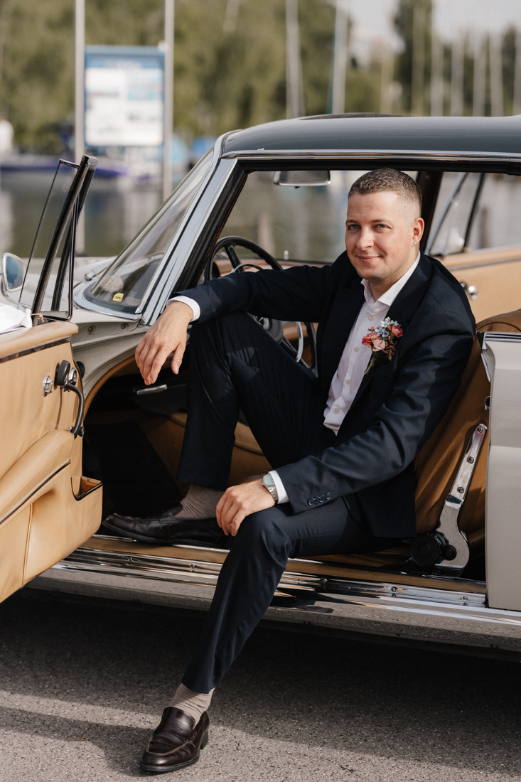 Mann im Anzug sitzt in Oldtimer mit offenstehender Tür und Blumenschmuck