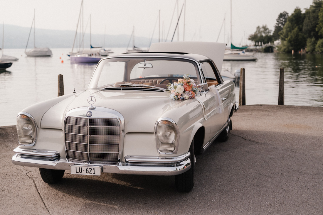 Ein weisser Oldtimer mit Blumenschmuck steht am Seeufer mit Segelbooten