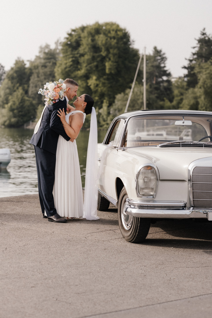 Umarmendes Paar mit Brautstrauss und Oldtimer Auto am Seeufer