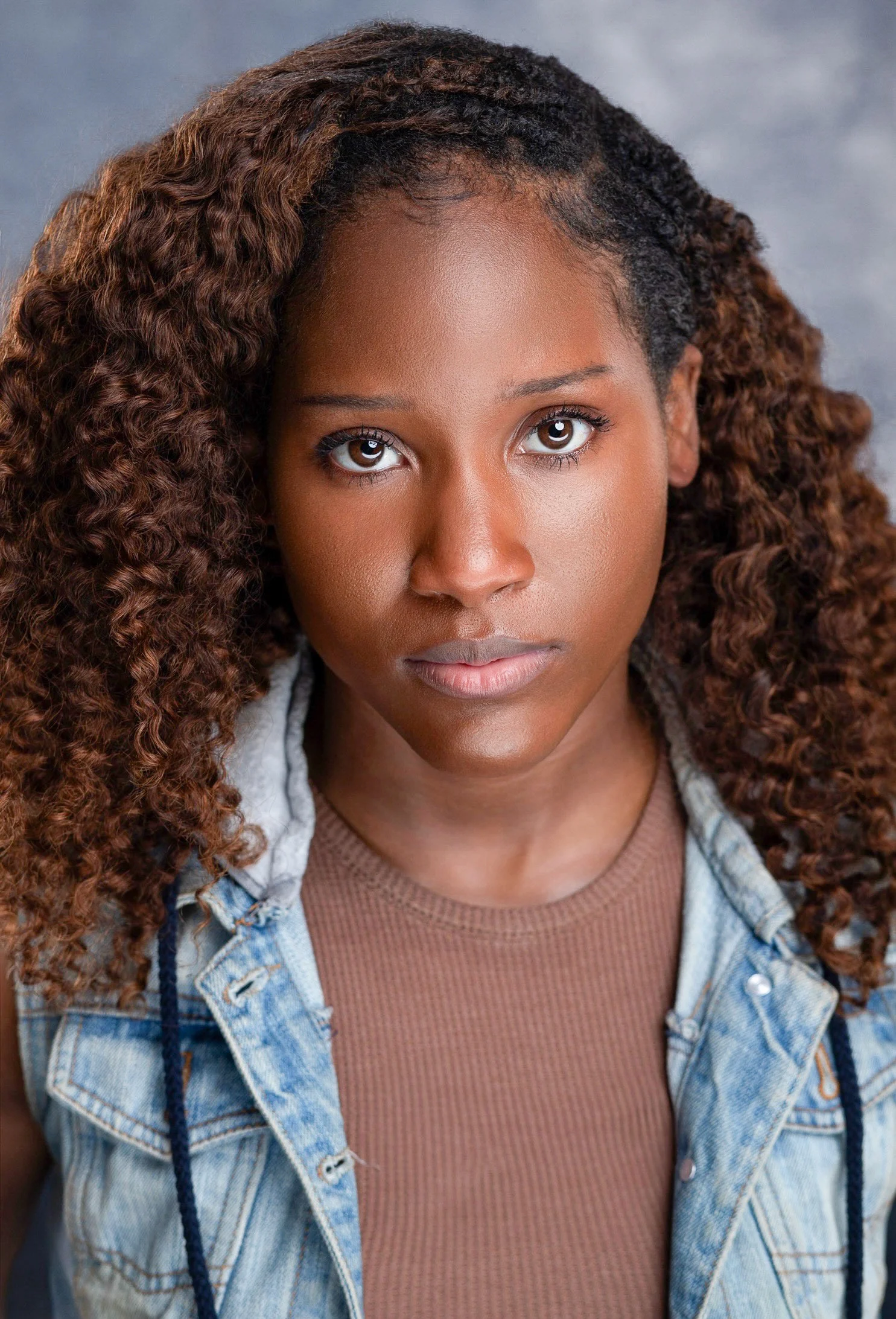 Marie Wilde with curly brown hair looking forward in a denim vest over a brown top against a gray background