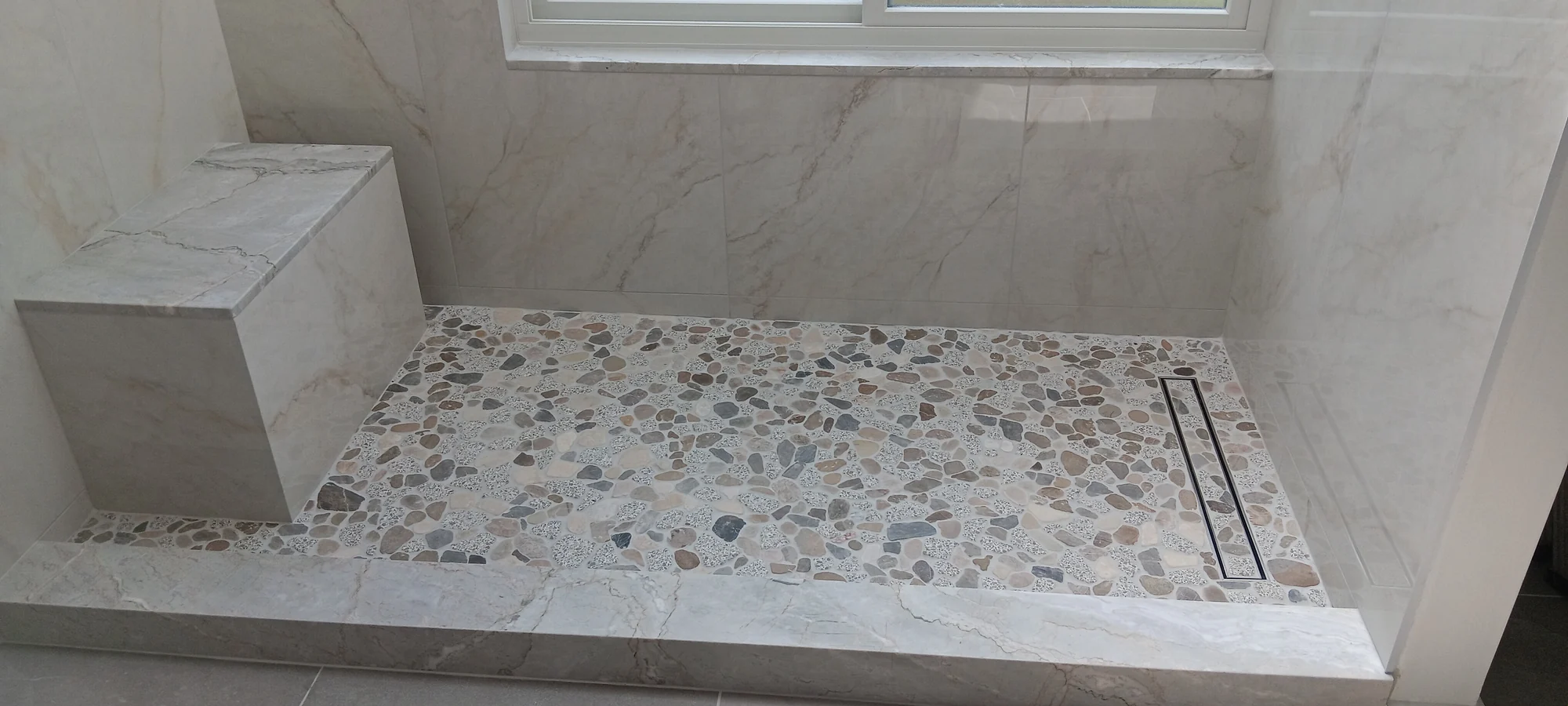 Shower tile detail with a built-in bench, pebble mosaic floor, and linear drain.