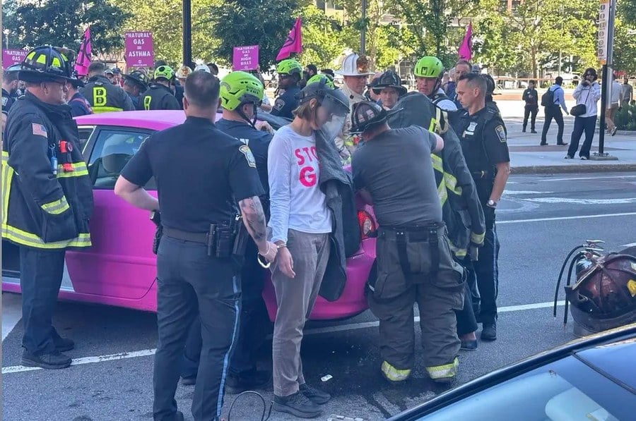A climate activist in a STOP GAS shirt is handcuffed by Boston Police after welding herself to a pink protest car