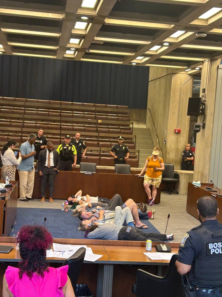 Boston City Council members and a Boston Police officer near the chamber floor while activists lay in a die-in
