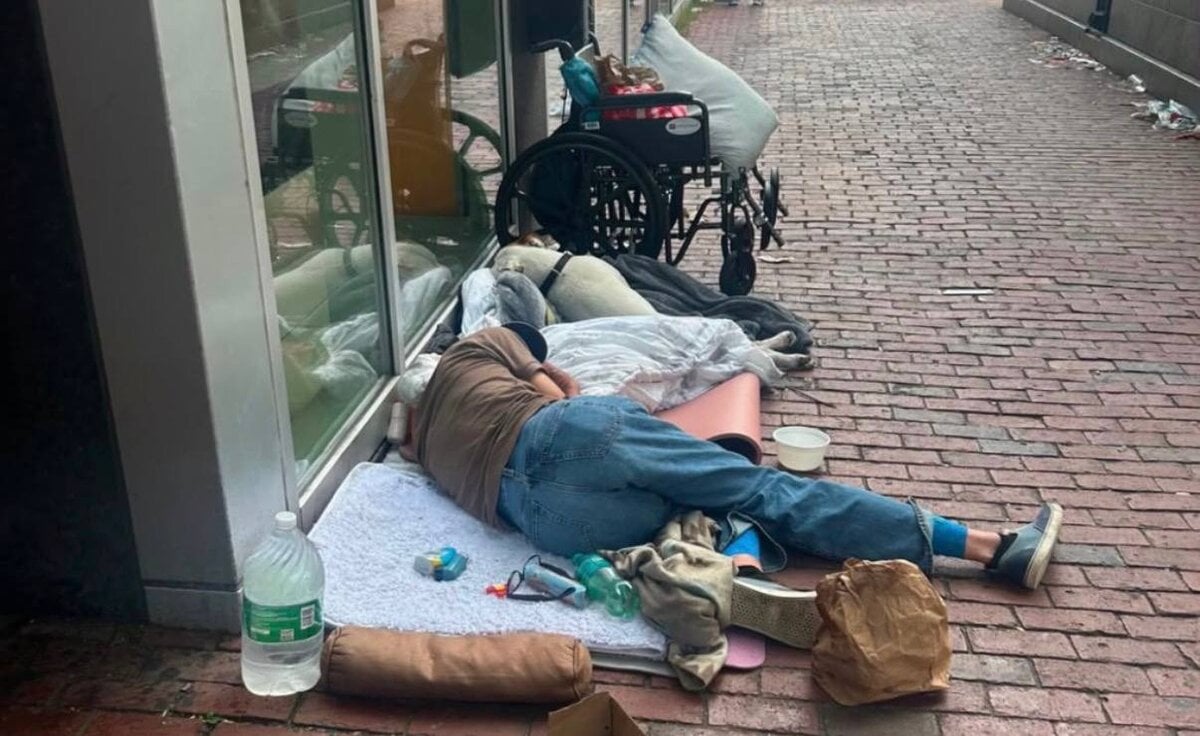 A person passed out on the sidewalk outside a Boylston Street storefront in Boston's Back Bay, with a wheelchair, bedding, and a cardboard donation box scattered on the pavement.