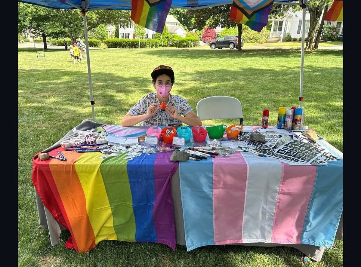 A young person seated at an OUT MetroWest activity table under a progress-pride flag canopy during a Pride event.
