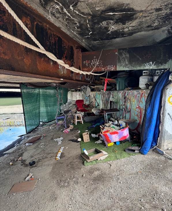 Inside the bridge: makeshift chairs, trash, and graffiti fill the hollow under the Charlesgate East off-ramp, where a hidden encampment has taken shape just feet from traffic above. Several propane tanks can be seen stored along the back wall.