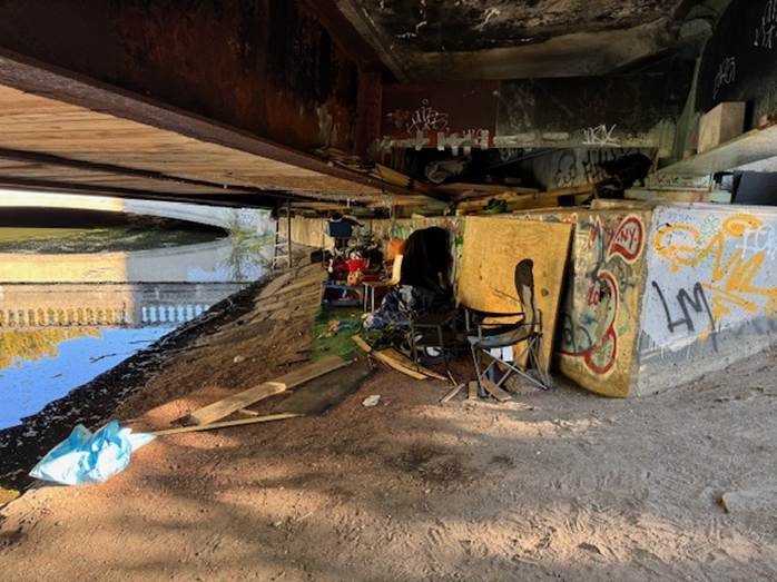 Beneath the Charlesgate East bridge, makeshift furniture and plywood walls form a hidden camp along the riverbank. What looks like a peaceful stretch of the Muddy River from above conceals a full-blown under-bridge settlement marked by graffiti, debris, and discarded supplies.