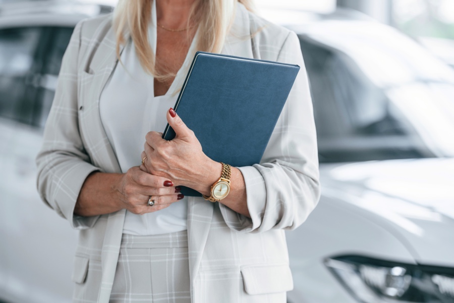 Conseillère automobile tenant un dossier devant un véhicule en concession, représentant l’immatriculation et les documents requis lors d’un achat de voiture usagée au Québec.