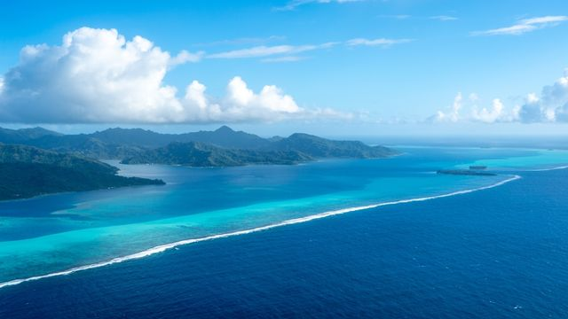 L'île de Tahaa, en Polynésie française, vue d'en haut, révèle un lagon turquoise, un récif corallien et des montagnes luxuriantes. Les couleurs vives de l'océan contrastent avec le bleu profond du Pacifique.
