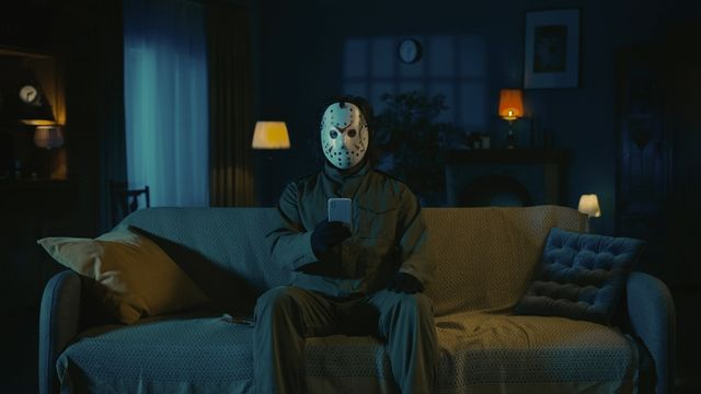 Jason Voorhees (Vendredi 13) assis sur le canapé du salon, smartphone en main, regardant les réseaux sociaux.