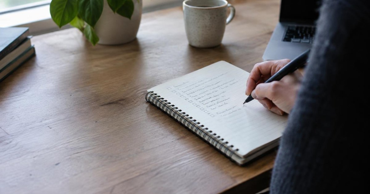 Hand writing on a checklist beside a laptop and coffee cup