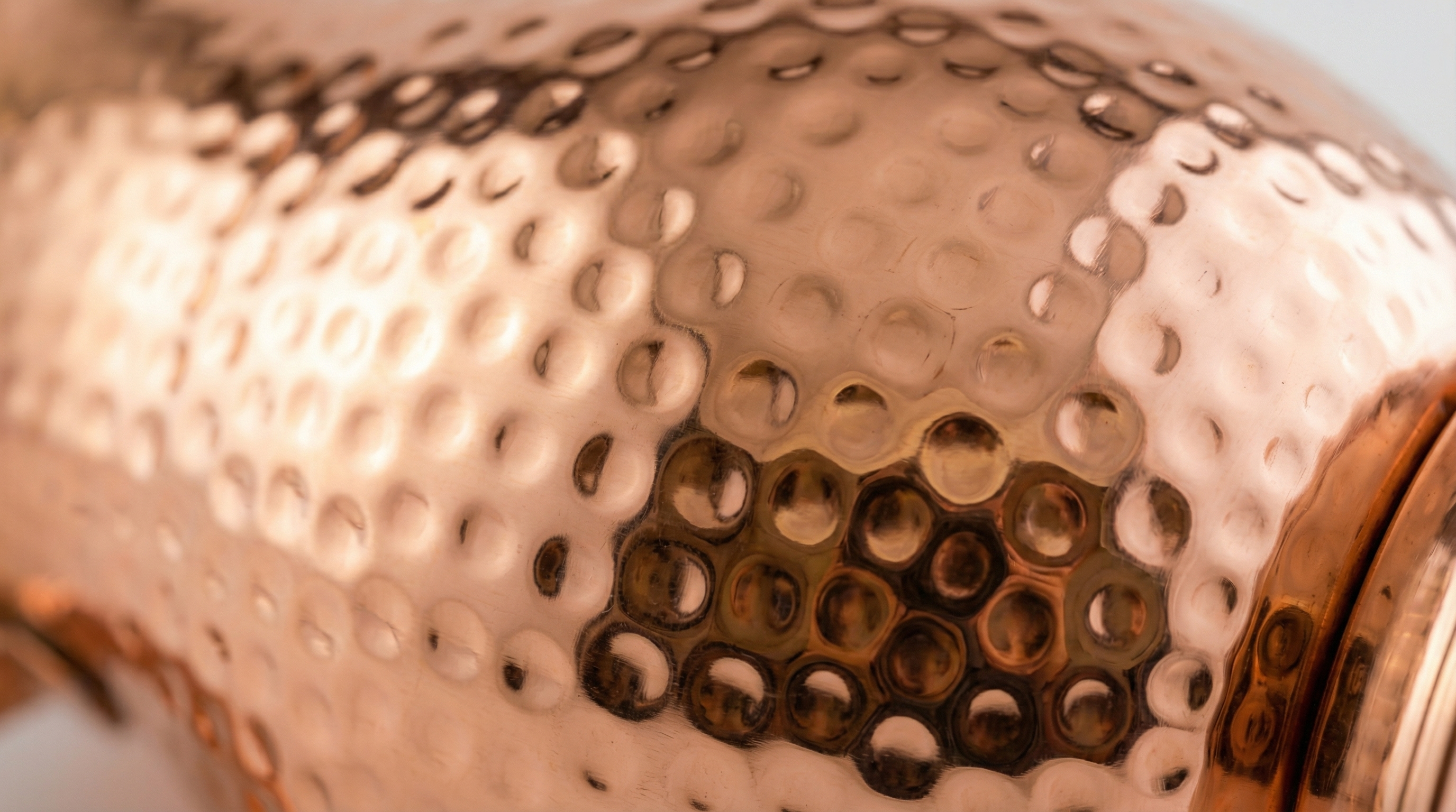 Extreme close-up macro shot of the side of a hammered copper vessel. The image details the individual hand-hammered dents and the warm, reflective, polished rose-gold surface. The lighting creates a beautiful play of light and shadow across the textured copper, emphasizing the artisan craftsmanship. Photorealistic, studio quality, 8k.