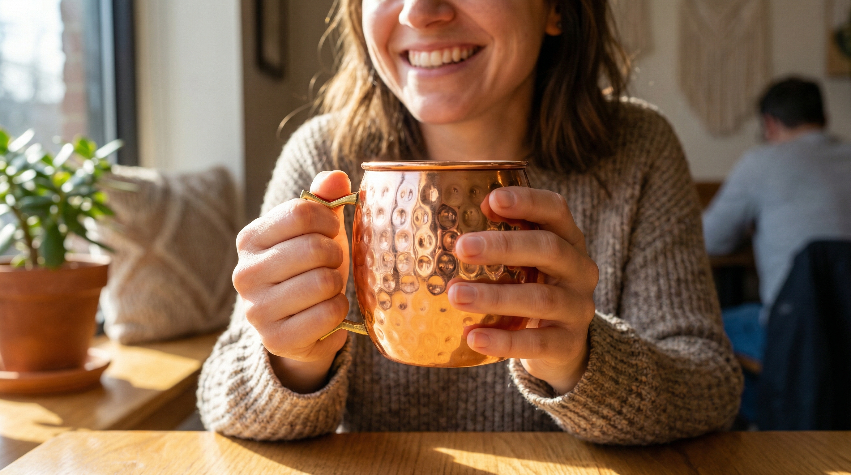 Close-up lifestyle shot of a happy woman in her 30s with a genuine smile, her hands wrapped around a hammered copper mug with an angular D-shaped brass handle. The focus is on the mug's texture and the warm interaction, set in a cozy, sunlit cafe. Photorealistic, candid lifestyle photography, 8k