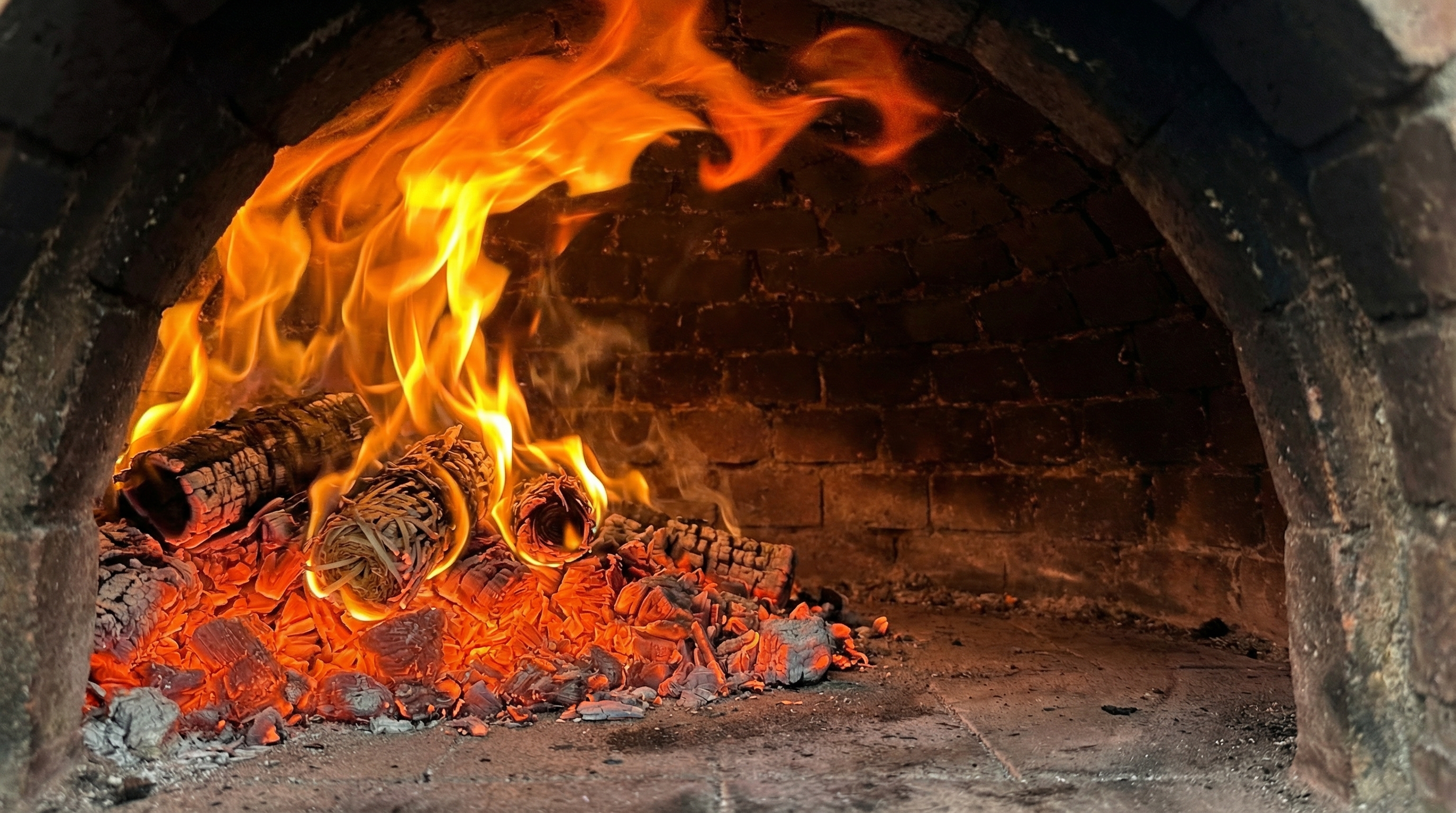 Interior shot of a pizza oven with a vibrant, active fire. Bright orange and yellow flames are licking the dome, and a deep bed of glowing red embers covers the floor on one side. The intense heat is palpable, creating a beautiful contrast between the fire and the dark, soot-stained brick. The scene is ready for cooking, conveying a sense of power, warmth, and anticipation. Photorealistic, macro detail, food photography, 8k.