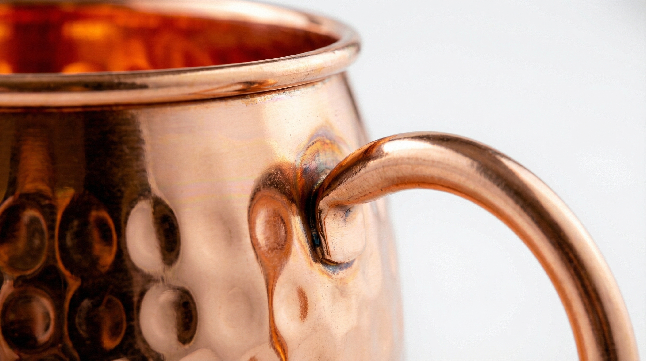 Extreme close-up macro shot of a real hammered copper mug, focusing on the point where the curved copper handle is expertly welded to the body. The image clearly shows the thickness of the copper at the rolled rim and the deep, authentic texture of the hammered dents. Clean, minimalist background with shallow depth of field, highlighting the craftsmanship, photorealistic, 8k.