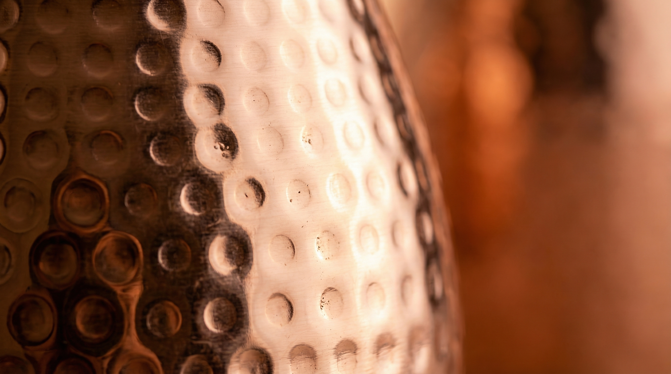 An extreme close-up, macro shot of the side of a hammered copper pitcher. The warm, natural light catches the individual, small circular dents of the hammered texture, creating a dynamic play of light and shadow. The focus is sharp on the craftsmanship and the rich, polished rose-gold copper color. The background is soft and out of focus, emphasizing the material's tactile beauty. Editorial photography style, shallow depth of field, 8k.