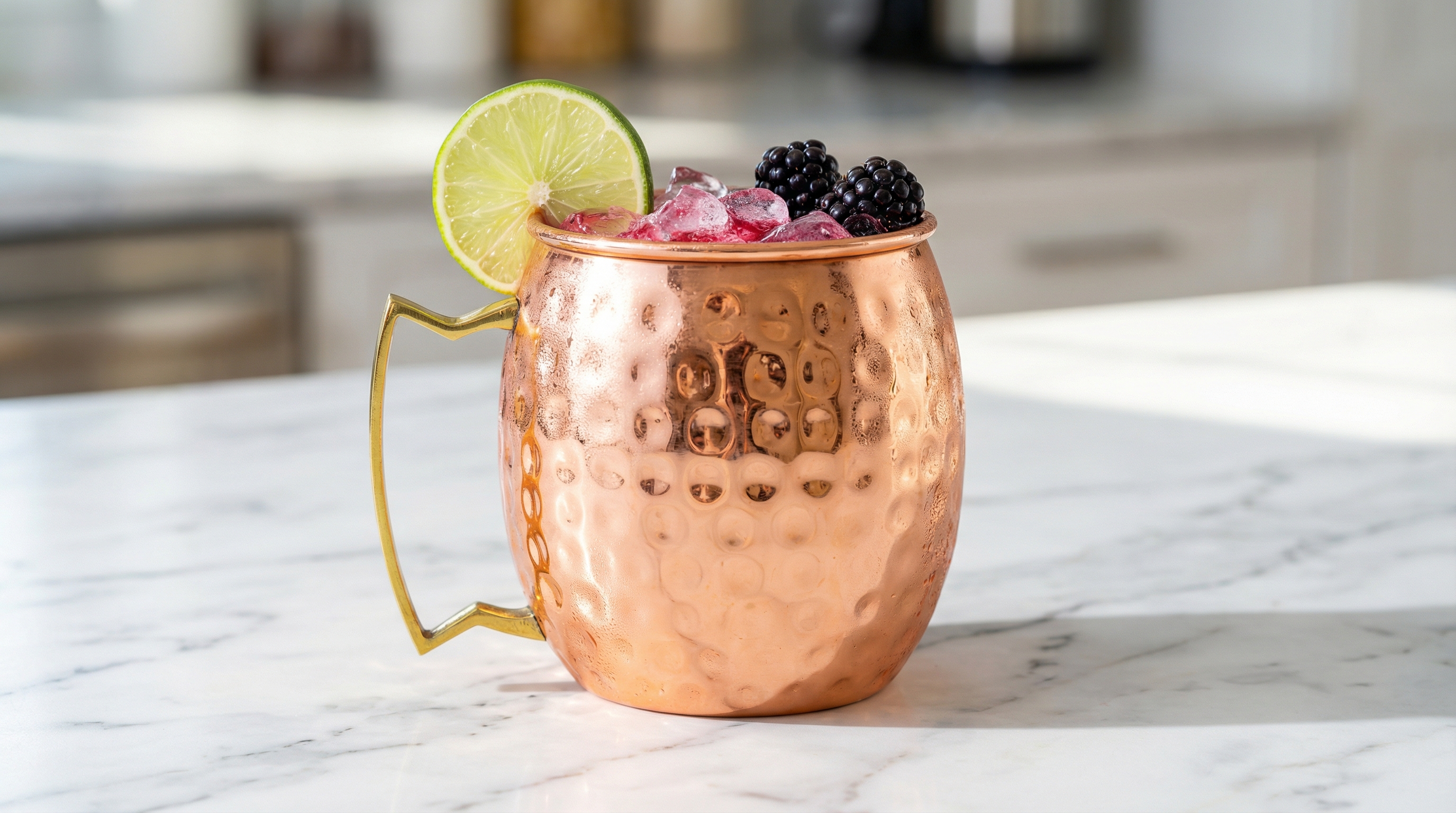 A vibrant, summery Mexican Mule served in a highly polished hammered copper mug with a distinct angular geometric brass handle. The cocktail has a bright pink hue from added raspberry or hibiscus, and is garnished with a lime wheel and fresh blackberries. The mug is set on a clean, white marble tabletop in bright, natural light, conveying a fresh and modern feel.