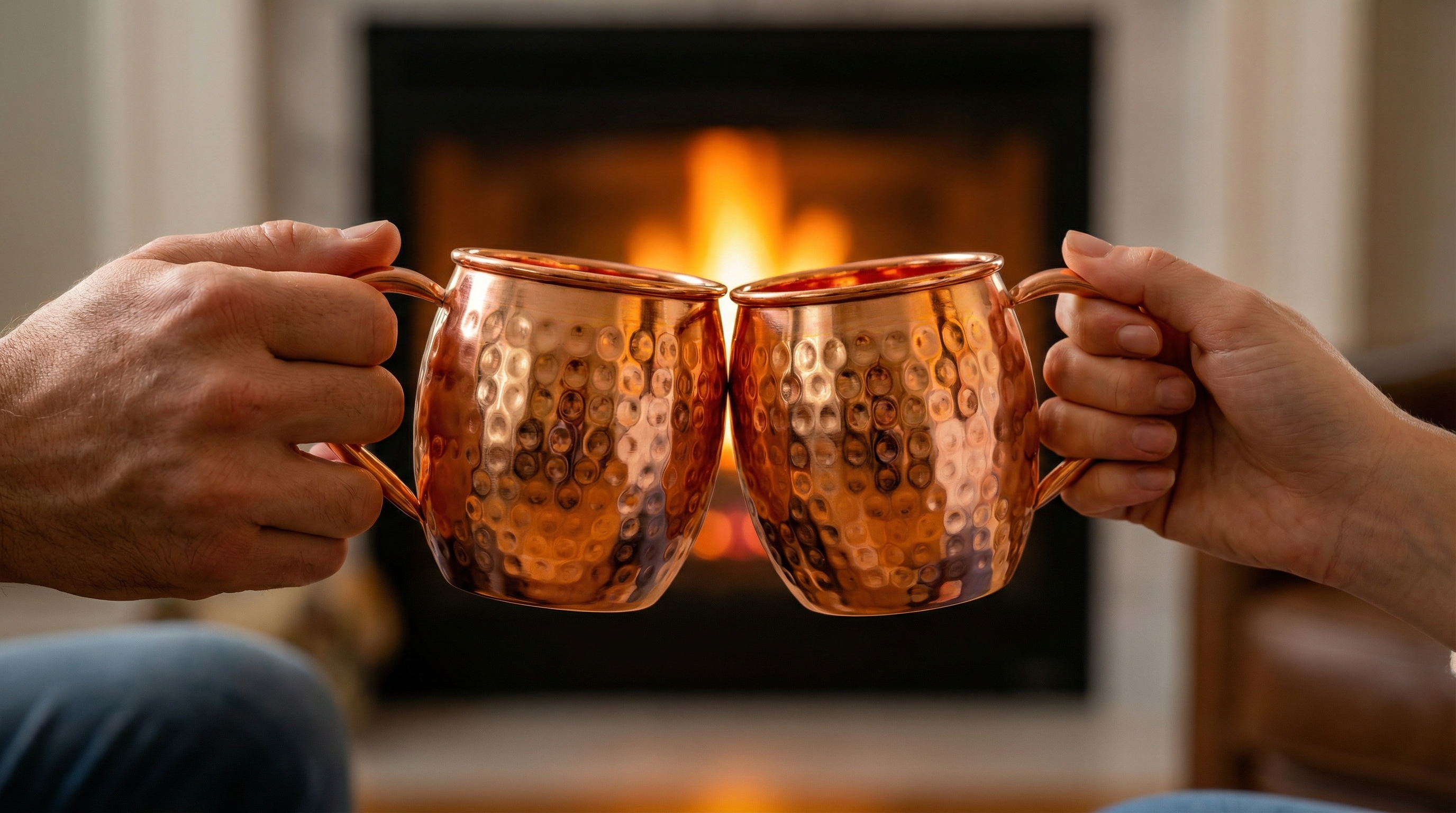 A close-up shot focusing on two highly polished hammered copper Moscow Mule mugs with welded, curved C-shaped pure copper handles clinking together in a toast. The hands holding them belong to a man and a woman. In the soft-focus background, the warm, orange glow of a fireplace is visible, adding to the romantic and cozy ambiance. Photorealistic, shallow depth of field, 8k