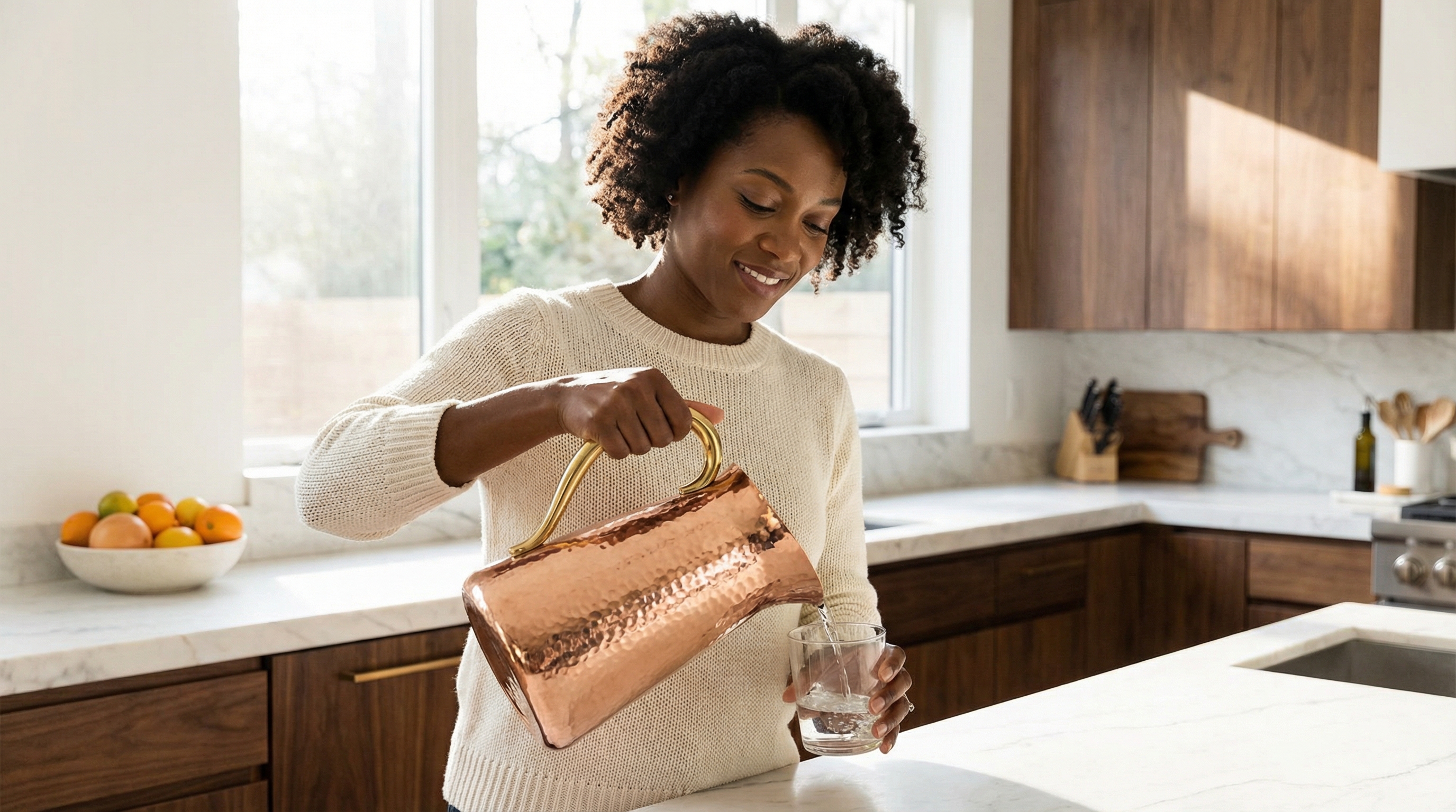 An elegant African American woman in her mid-30s, smiling warmly as she pours water from a stunning hammered copper water pitcher with a large, C-shaped polished brass handle into a glass. She is standing in a bright, minimalist modern kitchen with white marble countertops and dark wood cabinets. The scene is filled with soft, natural morning light, creating a feeling of serene luxury. Photorealistic, magazine editorial quality, 8k.