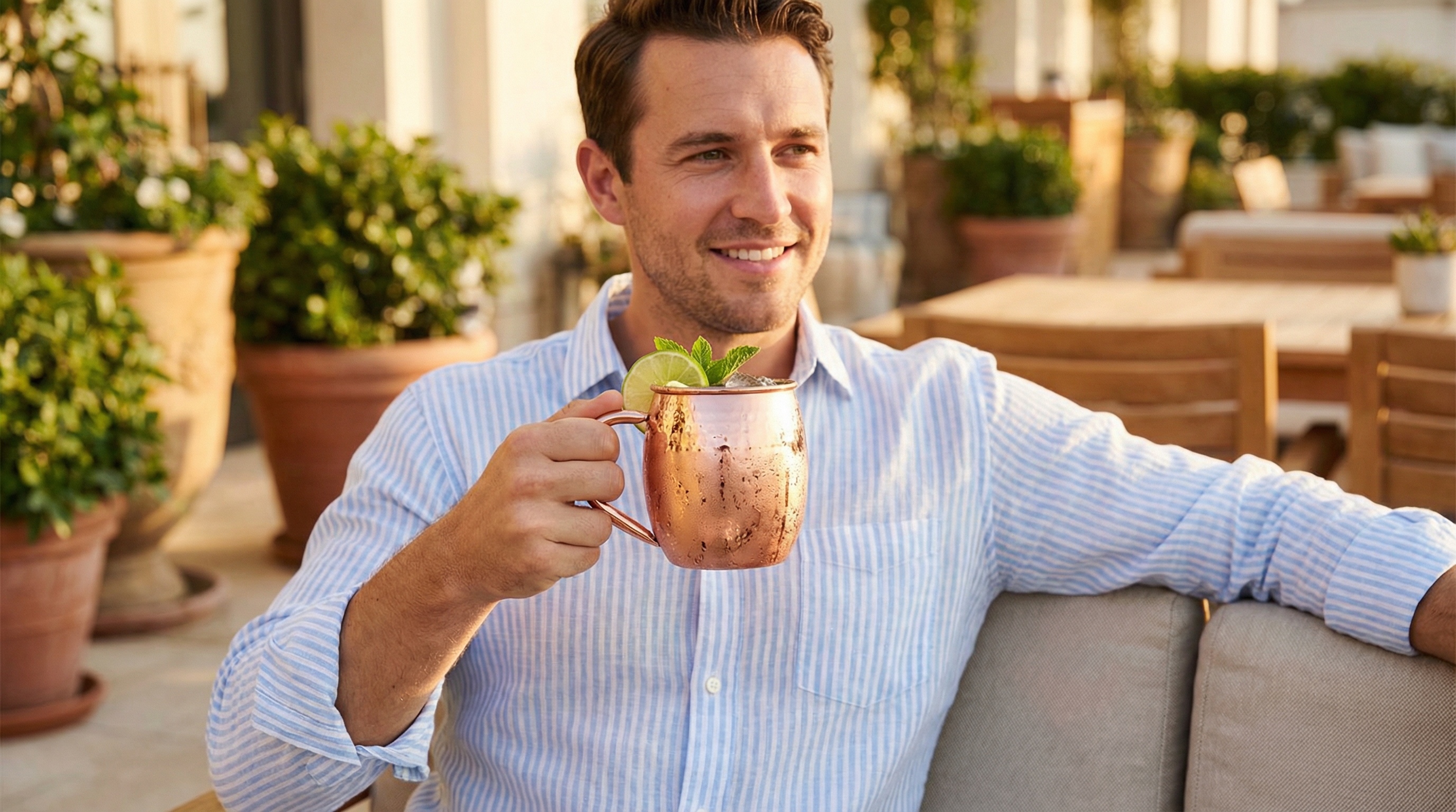 A handsome caucasian man in his early 30s, dressed in a fashionable light blue and white linen shirt, is enjoying a perfectly chilled Moscow Mule on a sunny, luxurious patio. He holds a highly polished, barrel-shaped Moscow Mule mug made of hammered pure copper with a distinct, elegant C-shaped copper handle welded seamlessly to the body, reflecting a bright rose-gold color. He has a relaxed, content smile, embodying a sense of sophisticated leisure. The background shows lush greenery and comfortable outdoor furniture, all bathed in warm, golden hour light. The image is photorealistic, with a shallow depth of field, capturing the magazine editorial quality, 8k.