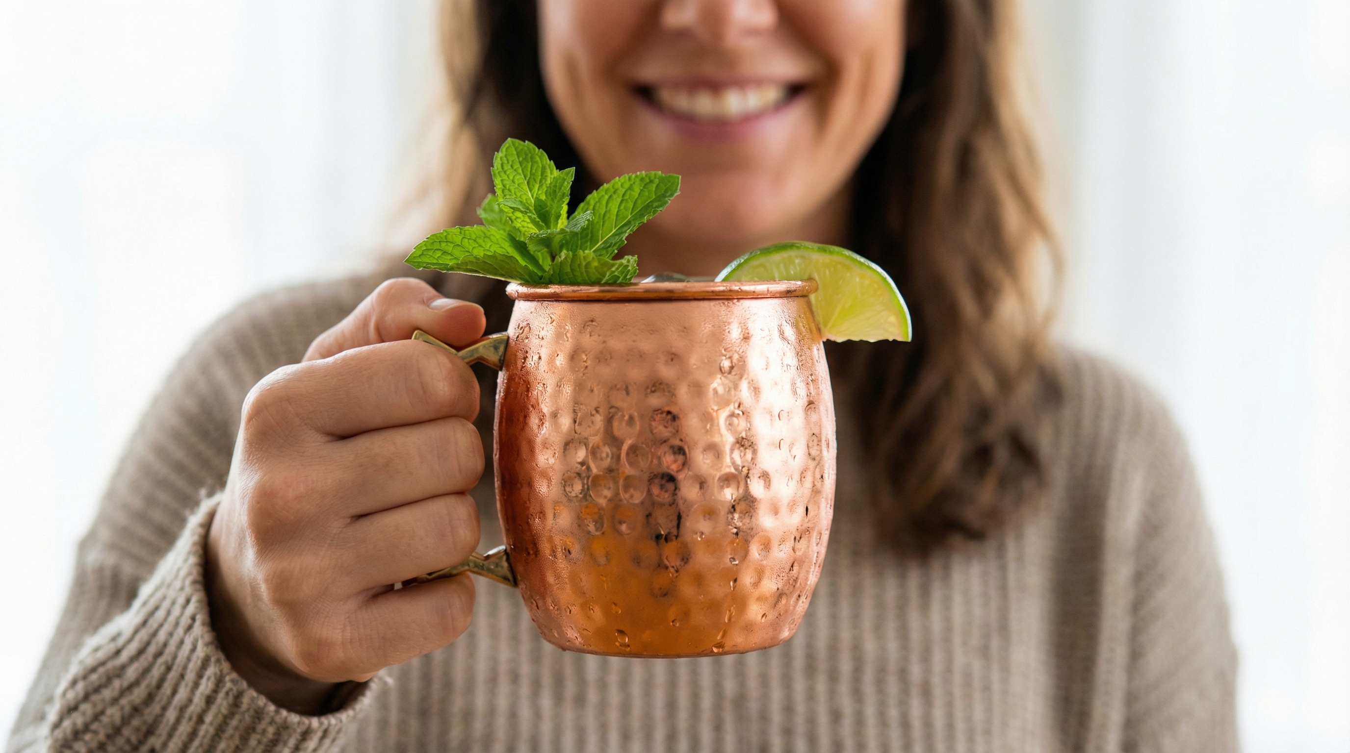 A close-up, artful shot of a refreshing Moscow Mule, held by a woman with a genuine, happy smile visible in the soft-focus background. The focus is squarely on the highly polished, barrel-shaped Moscow Mule mug made of hammered pure copper, which is covered in glistening condensation. A sprig of fresh mint and a bright green lime wedge garnish the drink, providing a pop of color against the warm rose-gold copper. The lighting is bright and natural, creating a feeling of authentic, candid lifestyle photography, 8k.