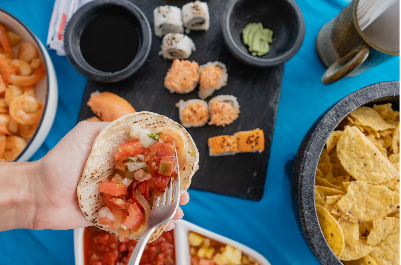 Hand holding a shrimp taco next to sushi rolls and chips on board