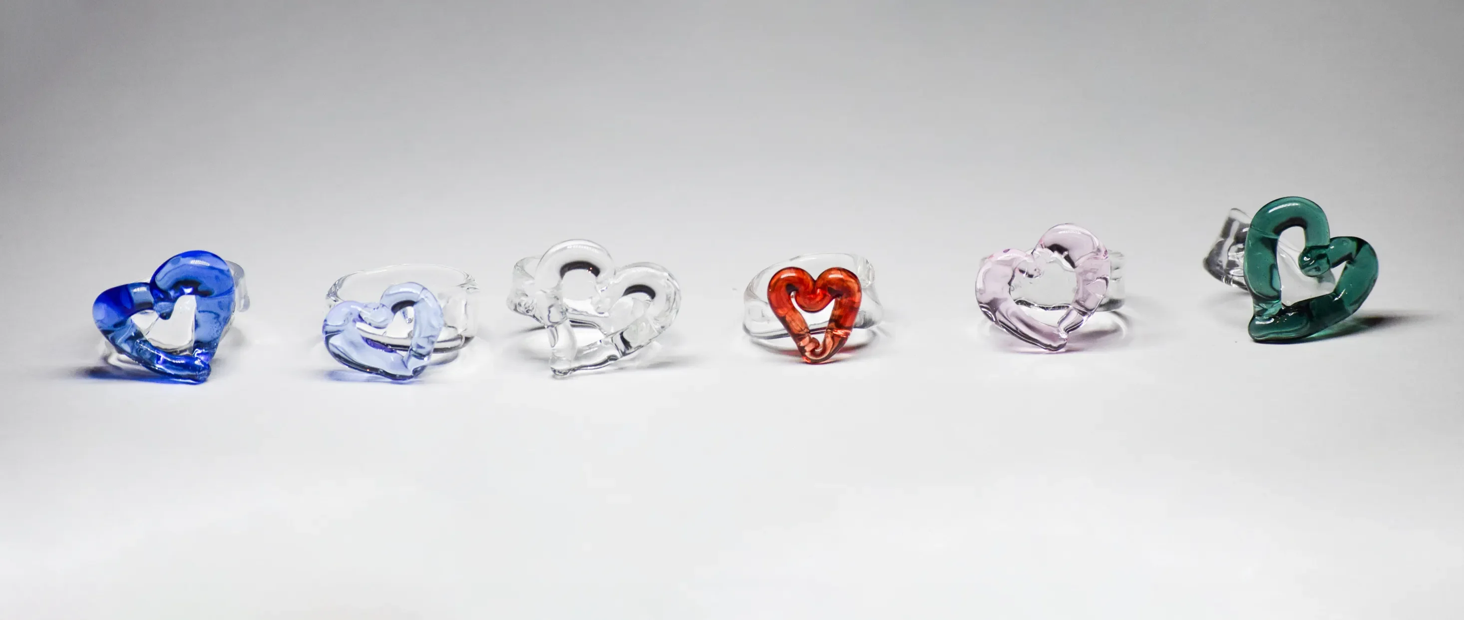 Seis anillos escultóricos de vidrio en forma de corazón dispuestos en fila, con colores de izquierda a derecha: azul cobalto, azul claro, cristal transparente, rojo con acento dorado, rosa pálido y verde esmeralda
