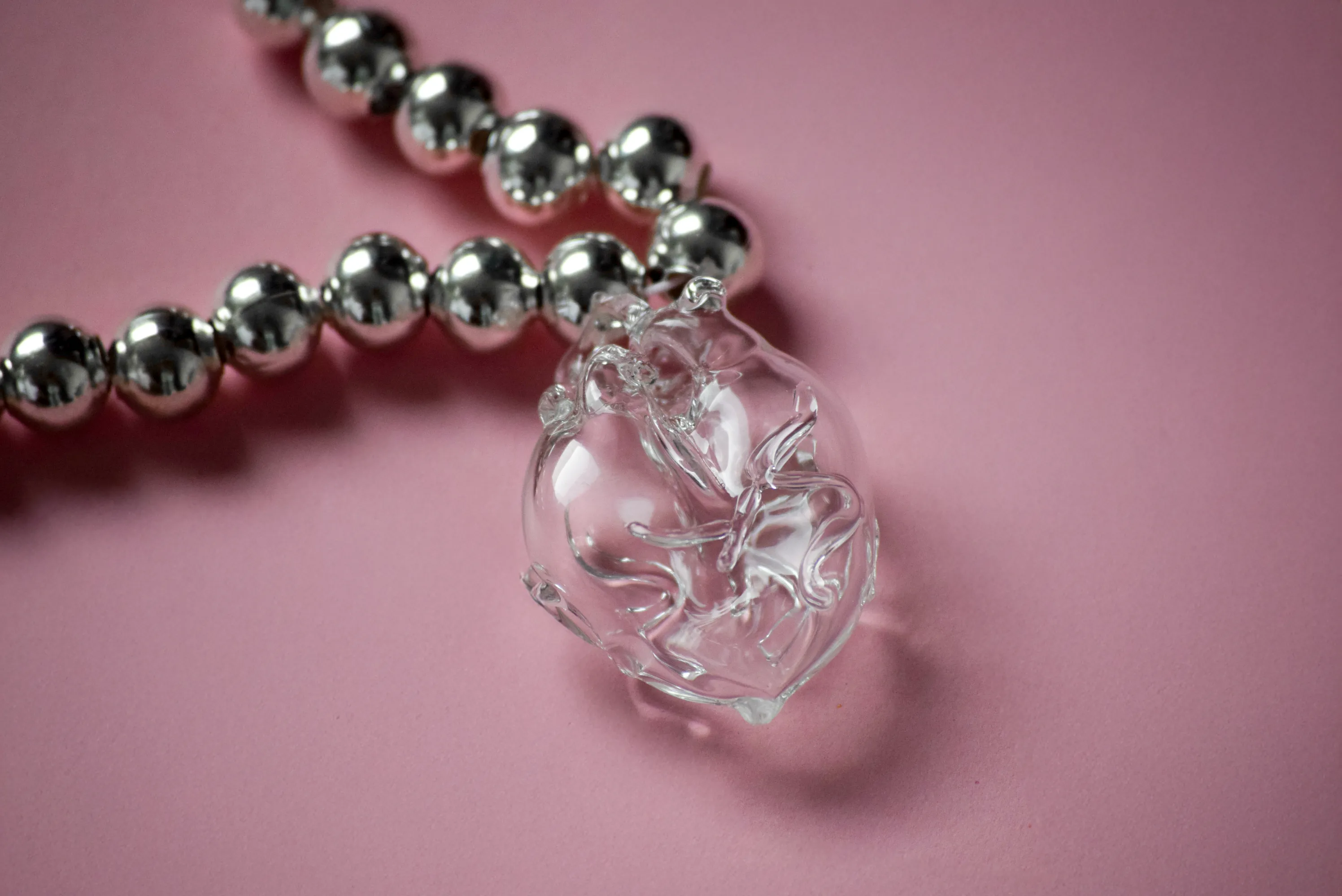 Close-up of a transparent glass anatomical heart pendant hanging from a silver ball chain necklace on a soft pink background
