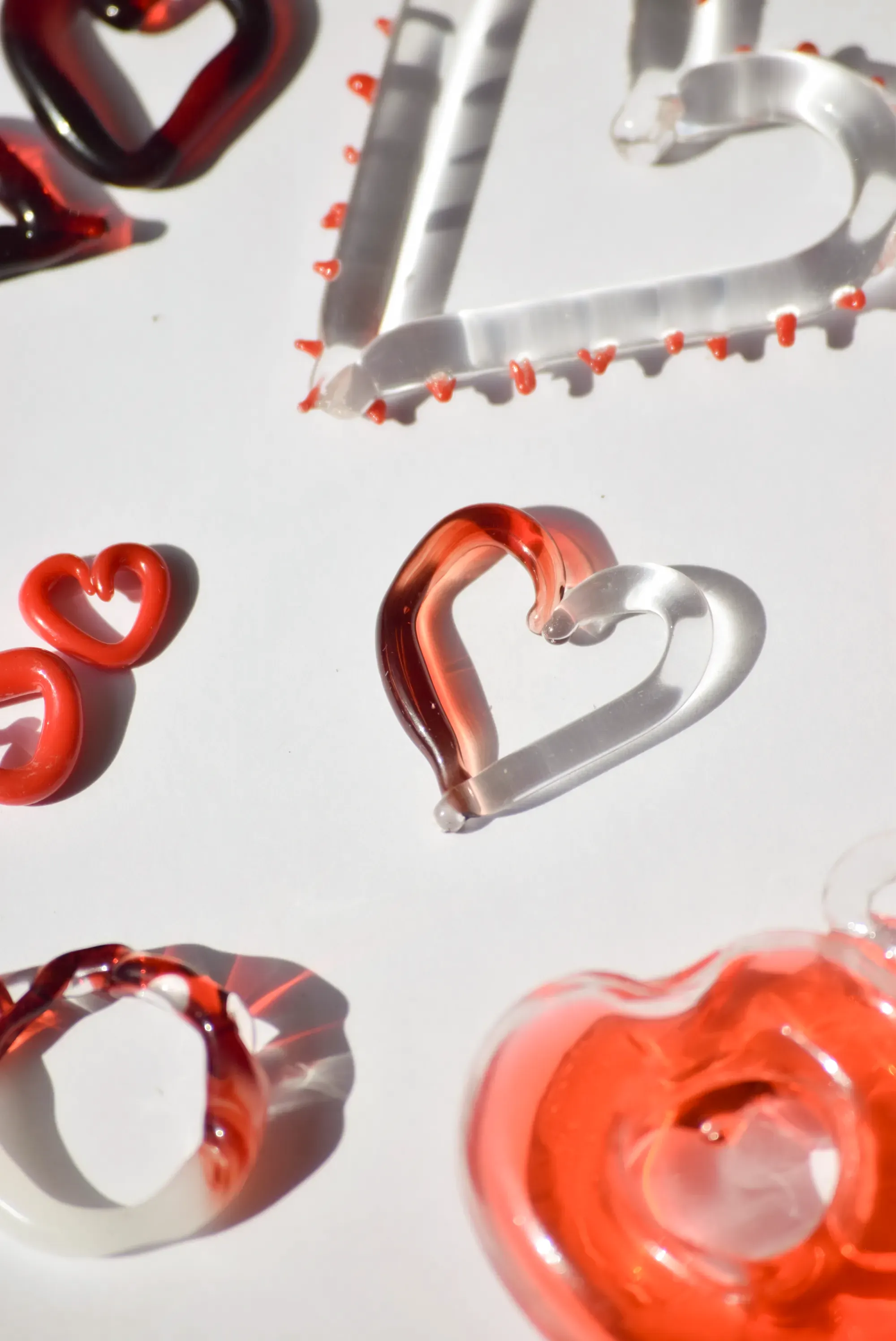 Composición en plano cenital de varias piezas de joyería de vidrio en forma de corazón sobre fondo blanco, con diseños degradados de rojo a transparente, anillos naranjas sólidos y detalles de cadena plateada con cuentas naranjasRetry
