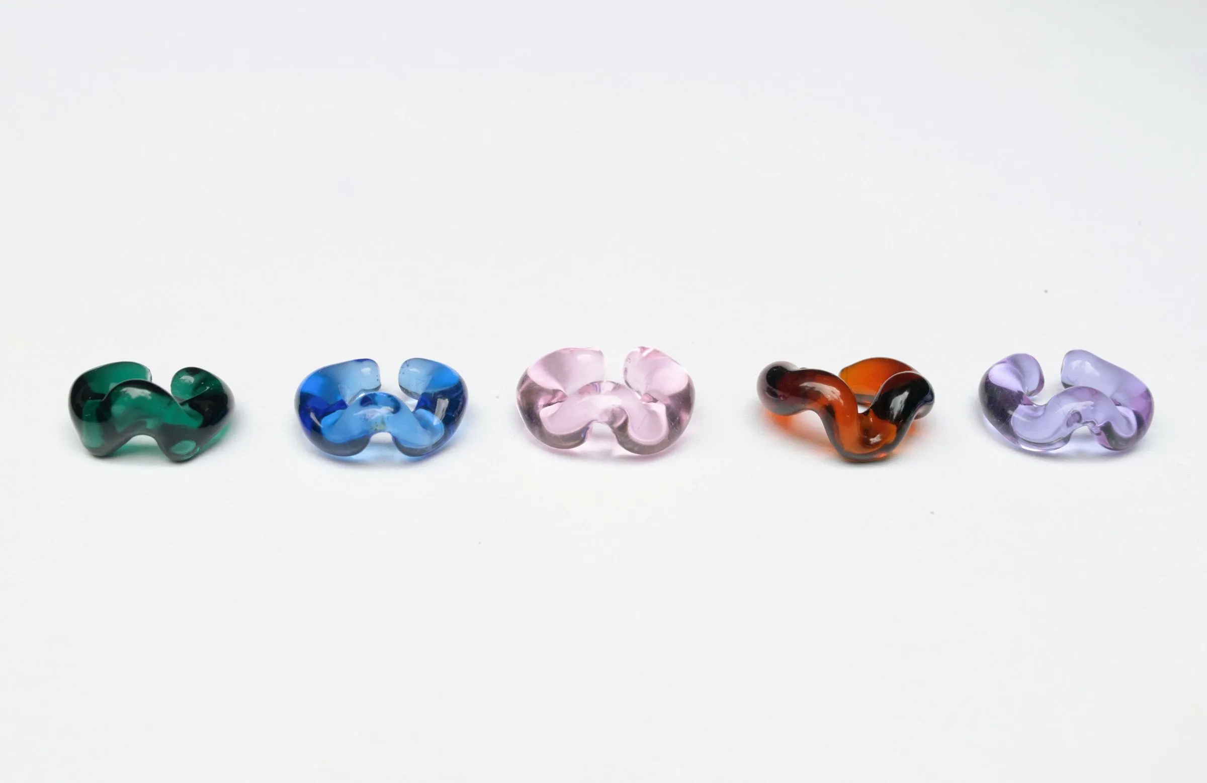 Five sculptural glass ear cuffs arranged in a row on white background, featuring organic flowing shapes in emerald green, cobalt blue, rose pink, amber-brown tortoiseshell, and lavender purple