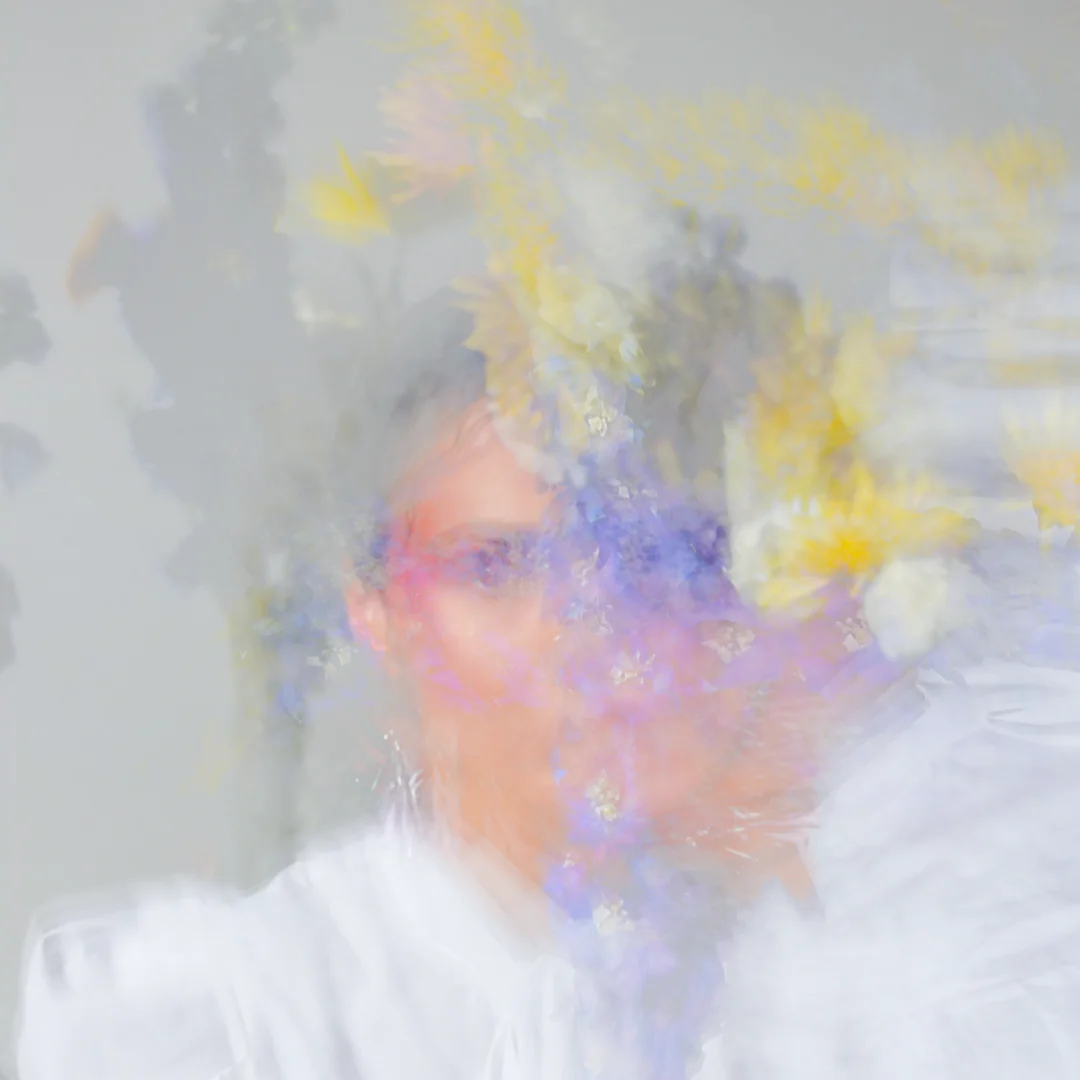 Person with artistic makeup surrounded by colorful powder explosion, including purple, yellow, and pink hues creating a dreamlike atmosphere