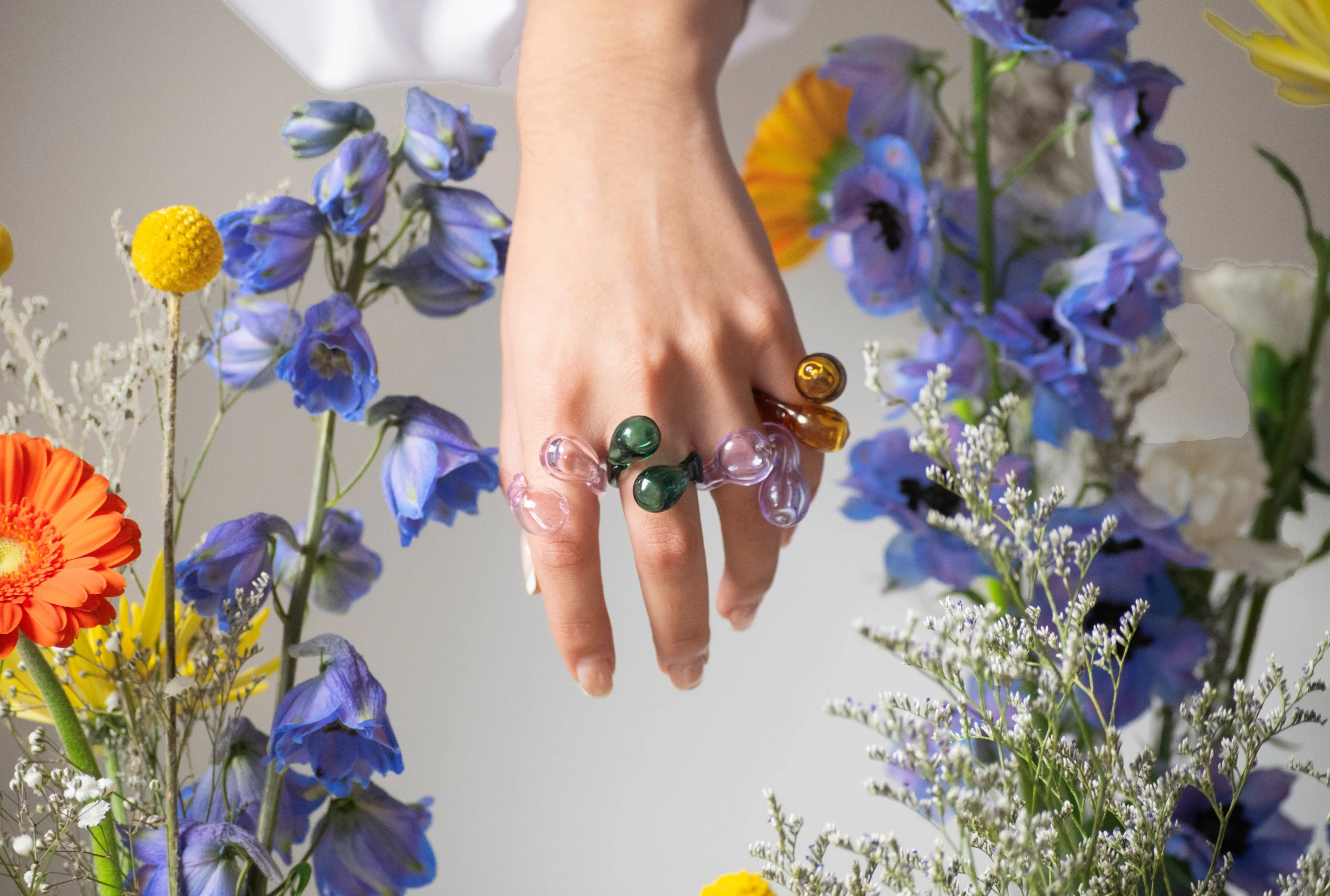 Hand displaying artistic glass rings with colorful spheres in pink, green, and amber tones, surrounded by blue delphiniums and orange gerbera daisies